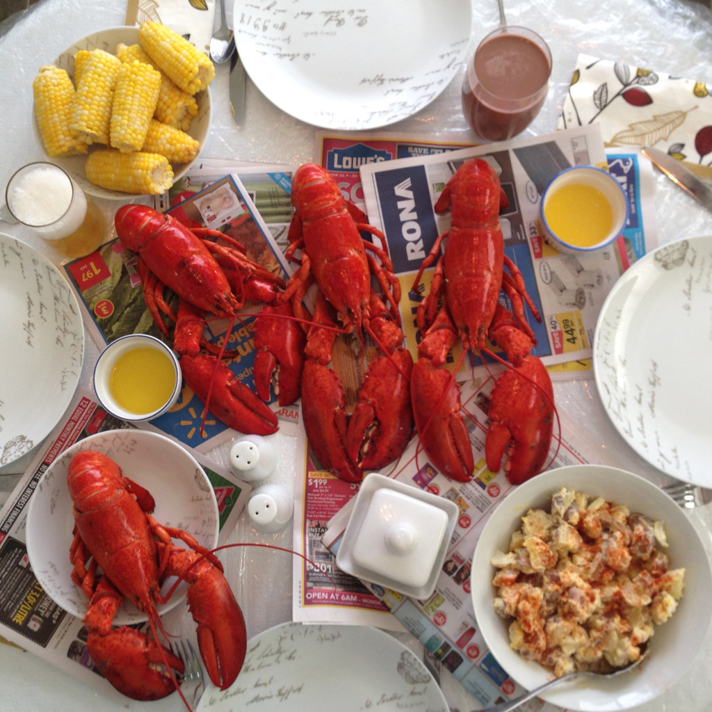 dinner table covered with newspaper and four lobsters, butter, corn and potato salad